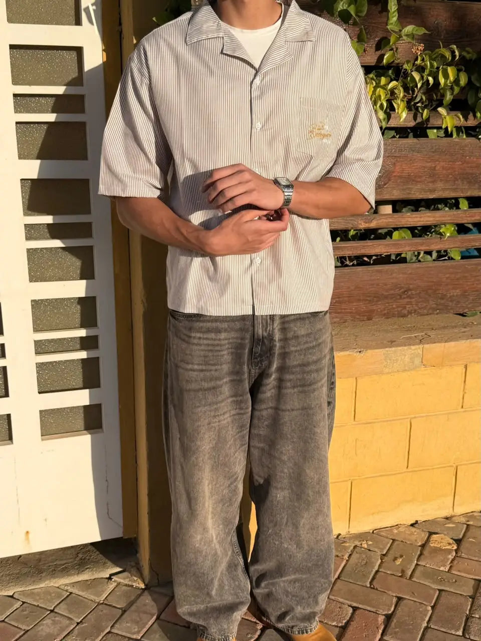 A person wearing a light beige short-sleeve button-up shirt with a small embroidered logo on the chest pocket, paired with faded gray jeans, standing outdoors against a wooden fence and brick wall.