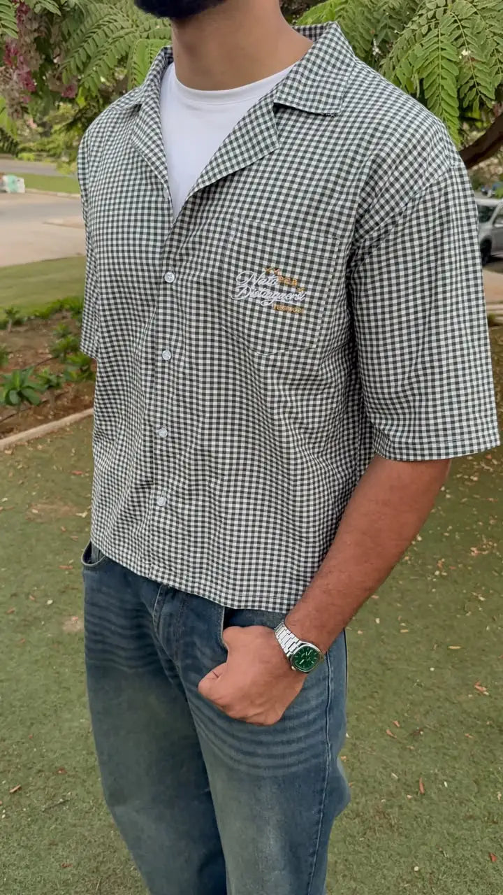 A man wearing a short-sleeve black and white gingham button-up shirt with a small embroidered logo on the left chest, paired with blue jeans, standing outdoors on a grassy lawn.