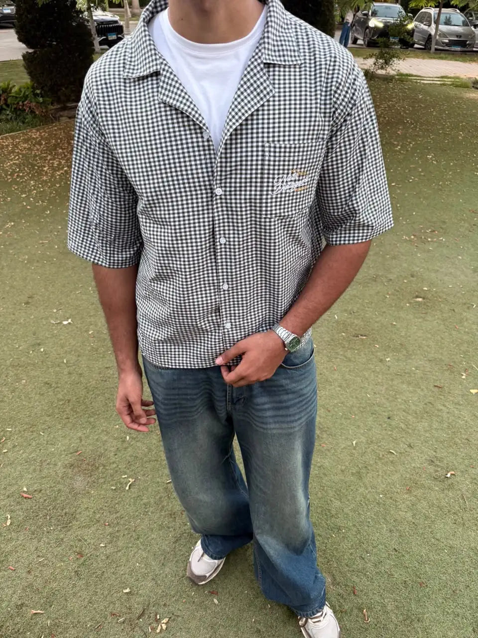 A man wearing a black and white gingham short-sleeve button-up shirt over a white t-shirt, paired with faded blue jeans and white sneakers, standing on an outdoor grassy lawn.